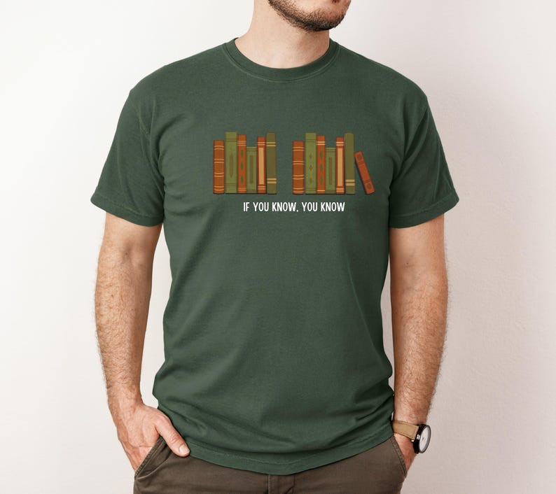May include: Dark green t-shirt featuring a row of stylized books in shades of green, orange, and brown. The text "IF YOU KNOW. YOU KNOW" is printed below the books. The shirt is a crew neck style.