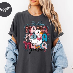 May include: Dark gray t-shirt featuring a cartoon duck with a pink bow, surrounded by the words "Mama Goose" in a colorful, patterned design. The shirt has short sleeves and a relaxed fit.