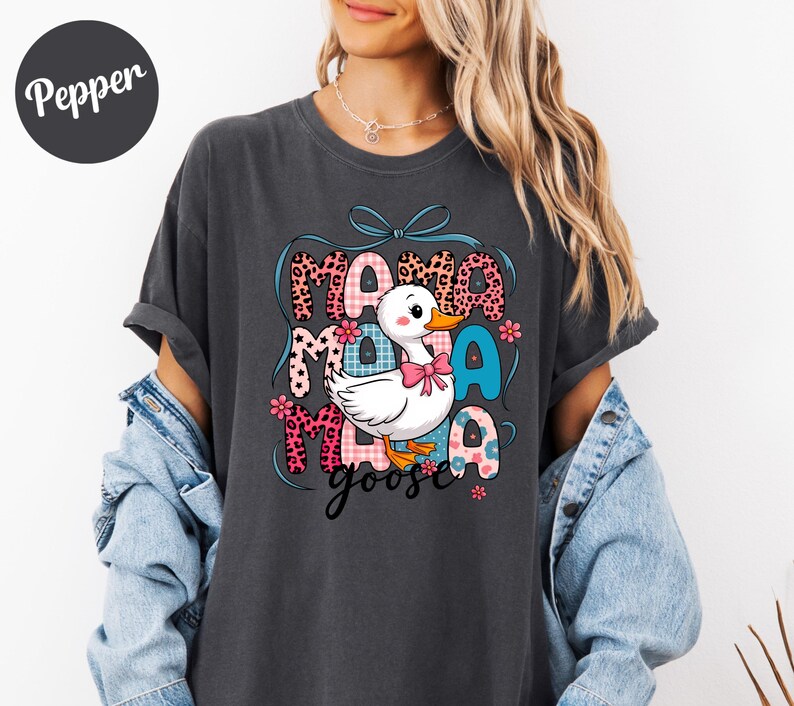 May include: Dark gray t-shirt featuring a cartoon duck with a pink bow, surrounded by the words "Mama Goose" in a colorful, patterned design. The shirt has short sleeves and a relaxed fit.