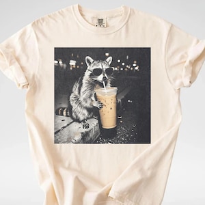 May include: Cream-colored t-shirt featuring a black and white graphic of a raccoon wearing sunglasses and drinking from a coffee cup. The raccoon is sitting on a ledge with a blurred background of city lights.
