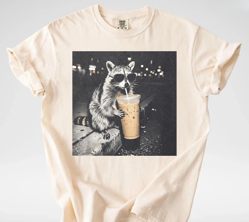 May include: Cream-colored t-shirt featuring a black and white graphic of a raccoon wearing sunglasses and drinking from a coffee cup. The raccoon is sitting on a ledge with a blurred background of city lights.