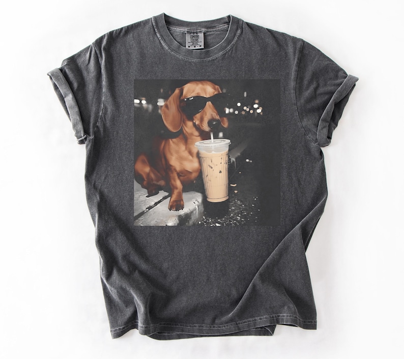 May include: Dark gray t-shirt featuring a graphic of a dachshund wearing sunglasses and drinking an iced coffee. The dog is brown with black sunglasses. The iced coffee is in a clear plastic cup with a white straw.