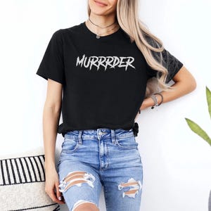 May include: Black t-shirt with the word "MURRRDER" in white, stylized, jagged lettering. The shirt is paired with blue denim jeans with distressed details. The model is wearing a necklace and bracelets.
