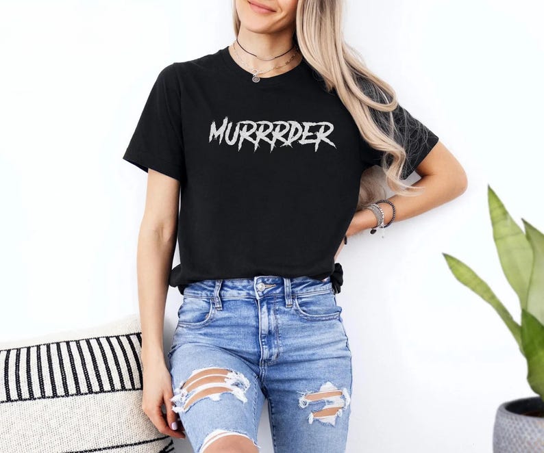 May include: Black t-shirt with the word "MURRRDER" in white, stylized, jagged lettering. The shirt is paired with blue denim jeans with distressed details. The model is wearing a necklace and bracelets.