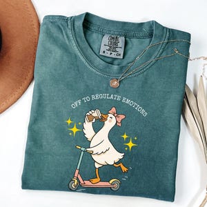 May include: A teal Comfort Colors t-shirt with a graphic of a goose riding a scooter and drinking coffee, with the text "OFF TO REGULATE EMOTIONS". A gold necklace is draped across the shirt.