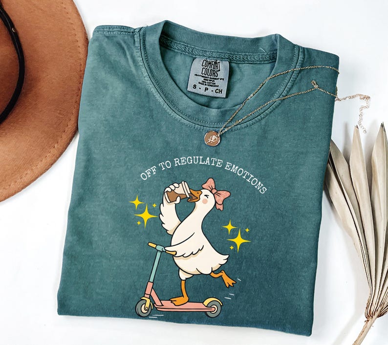 May include: A teal Comfort Colors t-shirt with a graphic of a goose riding a scooter and drinking coffee, with the text "OFF TO REGULATE EMOTIONS". A gold necklace is draped across the shirt.