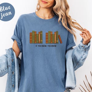 May include: Blue jean colored t-shirt with a graphic of a row of books in shades of brown and green. The text "IF YOU KNOW, YOU KNOW" is printed below the books. The shirt has short sleeves and a relaxed fit.
