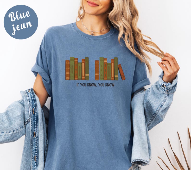 May include: Blue jean colored t-shirt with a graphic of a row of books in shades of brown and green. The text "IF YOU KNOW, YOU KNOW" is printed below the books. The shirt has short sleeves and a relaxed fit.