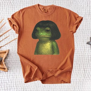 May include: A rust-colored t-shirt featuring a whimsical illustration of a green frog with a black bob haircut. The frog has large, expressive eyes and a serene expression. The shirt is made of soft material.
