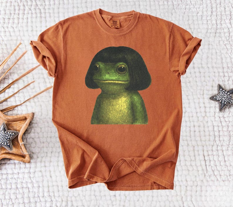 May include: A rust-colored t-shirt featuring a whimsical illustration of a green frog with a black bob haircut. The frog has large, expressive eyes and a serene expression. The shirt is made of soft material.