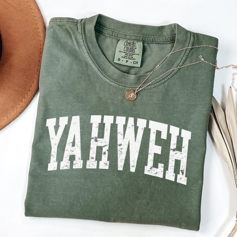 May include: Olive green t-shirt with the word "YAHWEH" in distressed white lettering. A gold-colored necklace with a pendant rests on the shirt. The shirt is labeled "Comfort Colors" and is size small.