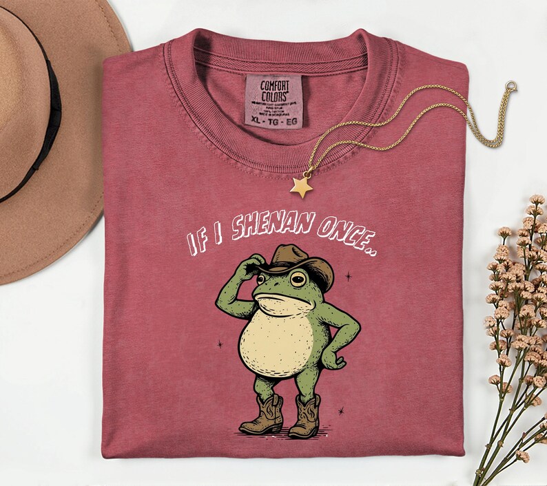 May include: A dusty rose Comfort Colors t-shirt featuring a cartoon frog in a cowboy hat and boots. The text "IF I SHENAN ONCE.." is above the frog. A gold star pendant necklace and a brown hat are also in the image.