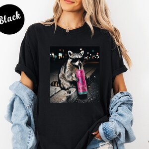May include: Black t-shirt featuring a graphic of a raccoon wearing sunglasses and drinking from a pink can. The raccoon is set against a blurred city night scene. The word "Black" is printed on a black circle.