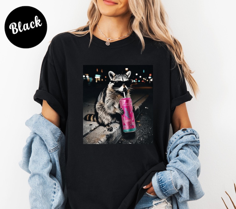 May include: Black t-shirt featuring a graphic of a raccoon wearing sunglasses and drinking from a pink can. The raccoon is set against a blurred city night scene. The word "Black" is printed on a black circle.