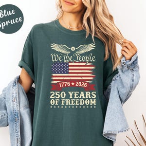 May include: Blue Spruce t-shirt with a graphic design featuring the American flag, the text "We the People," and "250 Years of Freedom" in white. The design includes the years 1776 and 2026. The shirt is a solid color.