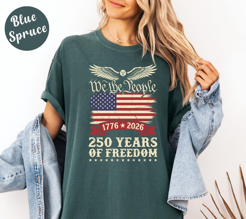 May include: Blue Spruce t-shirt with a graphic design featuring the American flag, the text "We the People," and "250 Years of Freedom" in white. The design includes the years 1776 and 2026. The shirt is a solid color.