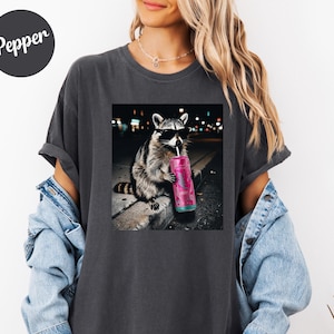May include: Dark gray t-shirt featuring a graphic of a raccoon wearing sunglasses and drinking from a pink can with a straw. The raccoon is sitting on a ledge with a city background. The word "Pepper" is on the upper left.