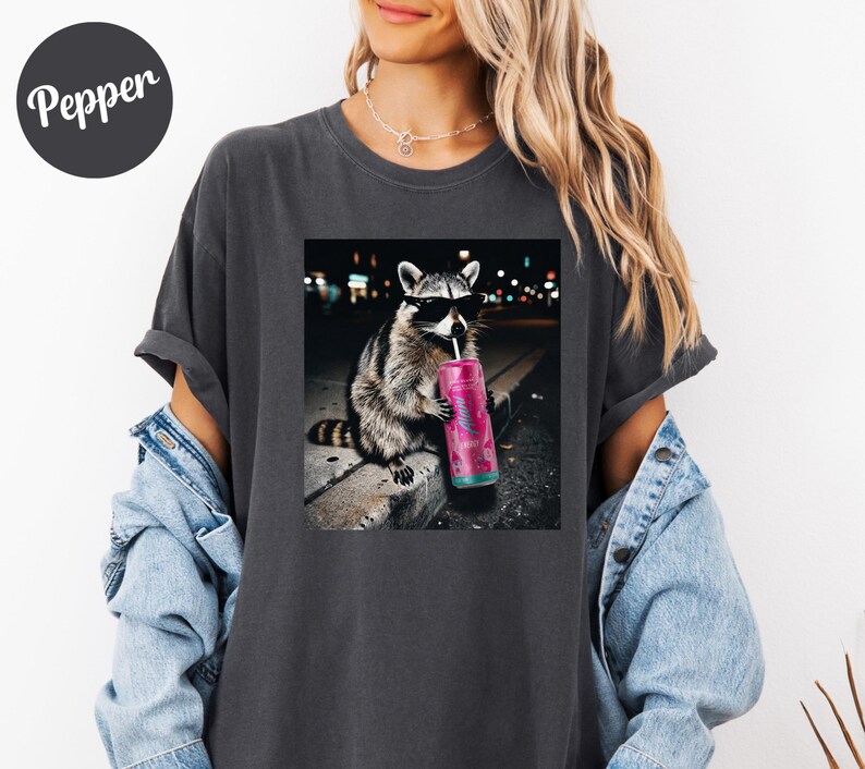 May include: Dark gray t-shirt featuring a graphic of a raccoon wearing sunglasses and drinking from a pink can with a straw. The raccoon is sitting on a ledge with a city background. The word "Pepper" is on the upper left.