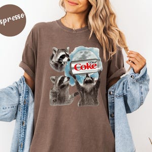 May include: Espresso-colored t-shirt featuring a graphic of three raccoons and a Diet Coke can against a blue and white moon background. The raccoons are in various poses, with one holding the soda can. The text "Diet Coke" is visible on the can.