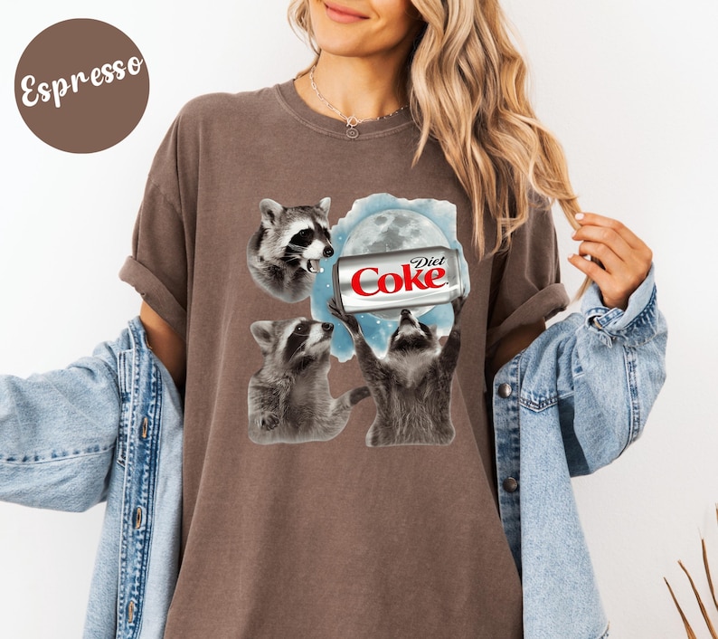 May include: Espresso-colored t-shirt featuring a graphic of three raccoons and a Diet Coke can against a blue and white moon background. The raccoons are in various poses, with one holding the soda can. The text "Diet Coke" is visible on the can.