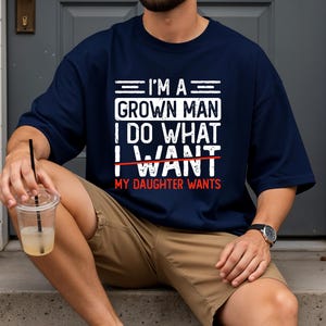 May include: Navy blue t-shirt with white text that reads "I'M A GROWN MAN I DO WHAT I WANT" with "WANT" crossed out and replaced with "DAUGHTER WANTS" in red. The shirt is worn by a person.