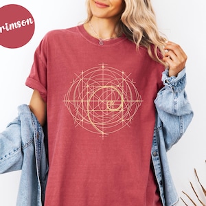 以下が含まれることがあります： クリムゾンカラーのTシャツ。金色の螺旋模様のデザイン。グラフィックは線と曲線で構成された幾何学模様です。シャツは半袖でゆったりとしたフィット感です。「Crimson」の文字が左上の円の中にあります。