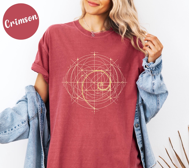 以下が含まれることがあります： クリムゾンカラーのTシャツ。金色の螺旋模様のデザイン。グラフィックは線と曲線で構成された幾何学模様です。シャツは半袖でゆったりとしたフィット感です。「Crimson」の文字が左上の円の中にあります。