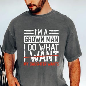 May include: Dark gray t-shirt with white text that reads "I'M A GROWN MAN I DO WHAT I WANT MY DAUGHTER WANTS" in a bold, blocky font. The word "WANT" is crossed out with a red line. The shirt has short sleeves.