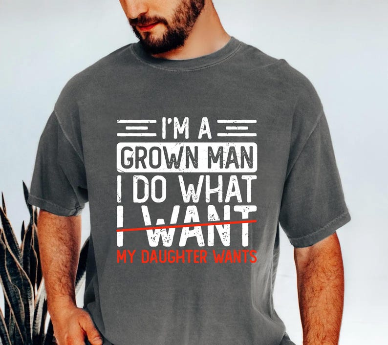 May include: Dark gray t-shirt with white text that reads "I'M A GROWN MAN I DO WHAT I WANT MY DAUGHTER WANTS" in a bold, blocky font. The word "WANT" is crossed out with a red line. The shirt has short sleeves.