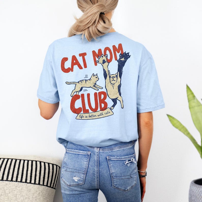 May include: Light blue t-shirt with a graphic that says "CAT MOM CLUB" in red and navy lettering. The graphic features two cartoon cats. One cat is running, and the other is reaching up. The text "life is better with cats" is below the word "CLUB".