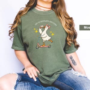 May include: Moss green t-shirt featuring a cartoon goose riding a scooter, holding a drink, and wearing a bow. The text "OFF TO REGULATE EMOTIONS" is above the goose. The shirt is a relaxed fit.