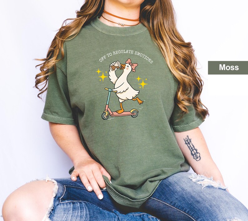 May include: Moss green t-shirt featuring a cartoon goose riding a scooter, holding a drink, and wearing a bow. The text "OFF TO REGULATE EMOTIONS" is above the goose. The shirt is a relaxed fit.
