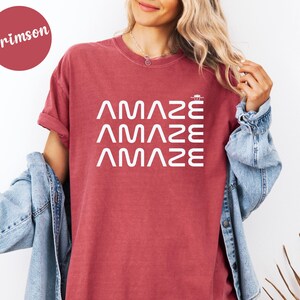 May include: Crimson-colored t-shirt with the word "AMAZE" printed three times in white. The shirt has short sleeves and a crew neck. The model is wearing a denim jacket.