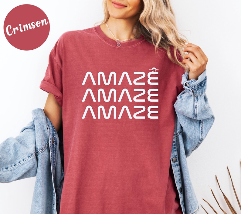 May include: Crimson-colored t-shirt with the word "AMAZE" printed three times in white. The shirt has short sleeves and a crew neck. The model is wearing a denim jacket.
