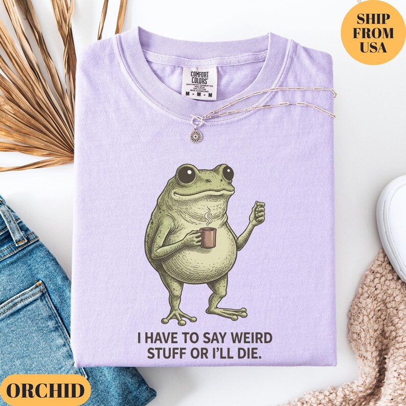 K&ouml;nnte beinhalten: Helllila T-Shirt mit einem Frosch, der eine Tasse h&auml;lt, und dem Text "I HAVE TO SAY WEIRD STUFF OR I'LL DIE." Eine silberne Halskette mit Sonnenanh&auml;nger liegt auf dem gefalteten Shirt.