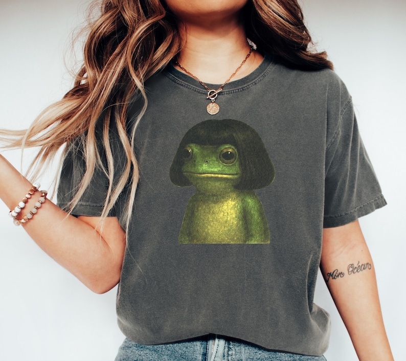 May include: Dark grey t-shirt featuring a green frog with a black wig. The frog has large eyes and a neutral expression. The shirt is a crew neck and has short sleeves. The overall style is casual and relaxed.
