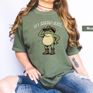 May include: A moss green t-shirt with a cartoon frog wearing a cowboy hat and boots. The frog is tipping its hat, with the text "If I Shenan Once..." above it. The shirt is a solid colour and has a relaxed fit.