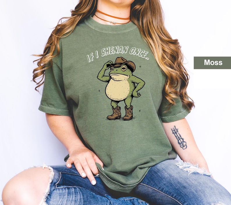 May include: A moss green t-shirt with a cartoon frog wearing a cowboy hat and boots. The frog is tipping its hat, with the text "If I Shenan Once..." above it. The shirt is a solid colour and has a relaxed fit.