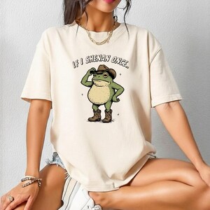May include: A cream-coloured t-shirt with a cartoon frog in a cowboy hat and boots. The frog is tipping its hat, with the text "IF I SHENAN ONCE.." above. The shirt has short sleeves and a relaxed fit.