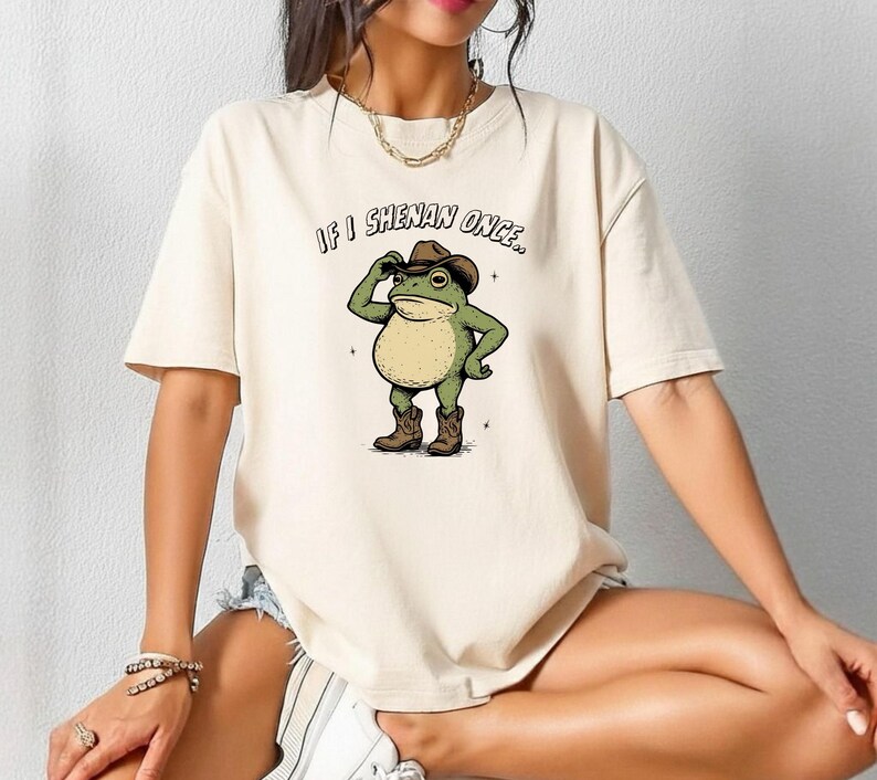 May include: A cream-coloured t-shirt with a cartoon frog in a cowboy hat and boots. The frog is tipping its hat, with the text "IF I SHENAN ONCE.." above. The shirt has short sleeves and a relaxed fit.
