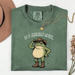 May include: Green Comfort Colors t-shirt featuring a cartoon frog in a cowboy hat and boots, with the text "IF I SHENAN ONCE..". A gold star necklace and a brown hat are also visible in the image.