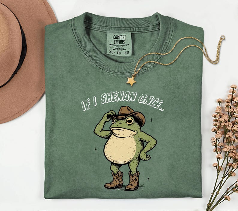 May include: Green Comfort Colors t-shirt featuring a cartoon frog in a cowboy hat and boots, with the text "IF I SHENAN ONCE..". A gold star necklace and a brown hat are also visible in the image.