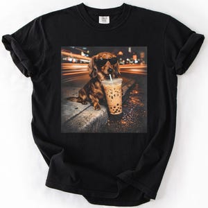 Puede incluir: Camiseta negra con un dise&ntilde;o de un perro salchicha con gafas de sol y un bubble tea. El perro marr&oacute;n est&aacute; sentado en una acera. El fondo muestra luces borrosas, sugiriendo una escena urbana.
