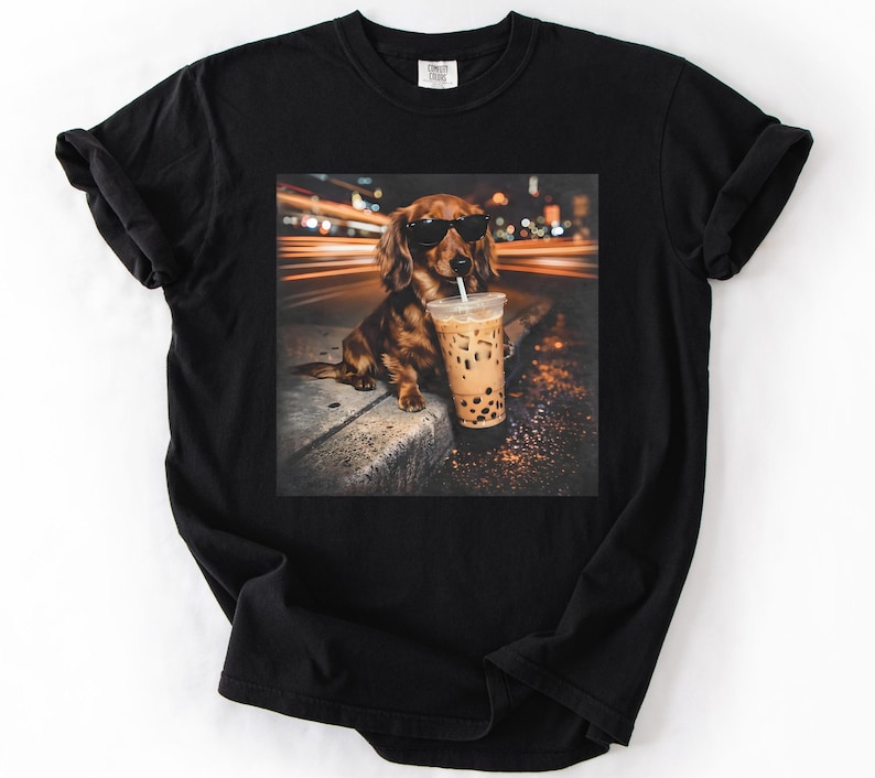 Puede incluir: Camiseta negra con un dise&ntilde;o de un perro salchicha con gafas de sol y un bubble tea. El perro marr&oacute;n est&aacute; sentado en una acera. El fondo muestra luces borrosas, sugiriendo una escena urbana.