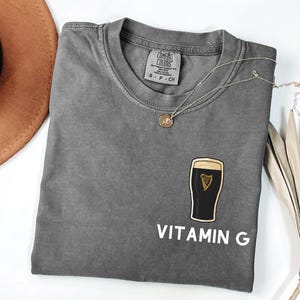 May include: A gray t-shirt with a crew neck and the text "VITAMIN G" in white. The shirt features a graphic of a pint of stout with a gold harp. A silver necklace with a small pendant is draped across the shirt.