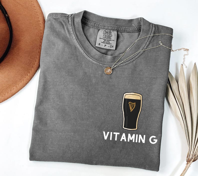 May include: A gray t-shirt with a crew neck and the text "VITAMIN G" in white. The shirt features a graphic of a pint of stout with a gold harp. A silver necklace with a small pendant is draped across the shirt.