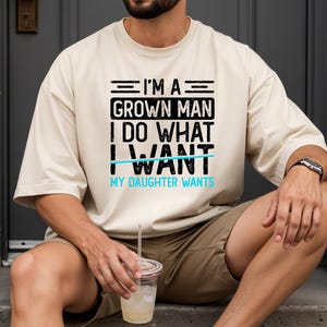 May include: A beige t-shirt with the text "I'M A GROWN MAN I DO WHAT I WANT MY DAUGHTER WANTS" in black and light blue. The person is wearing a watch and holding a drink.