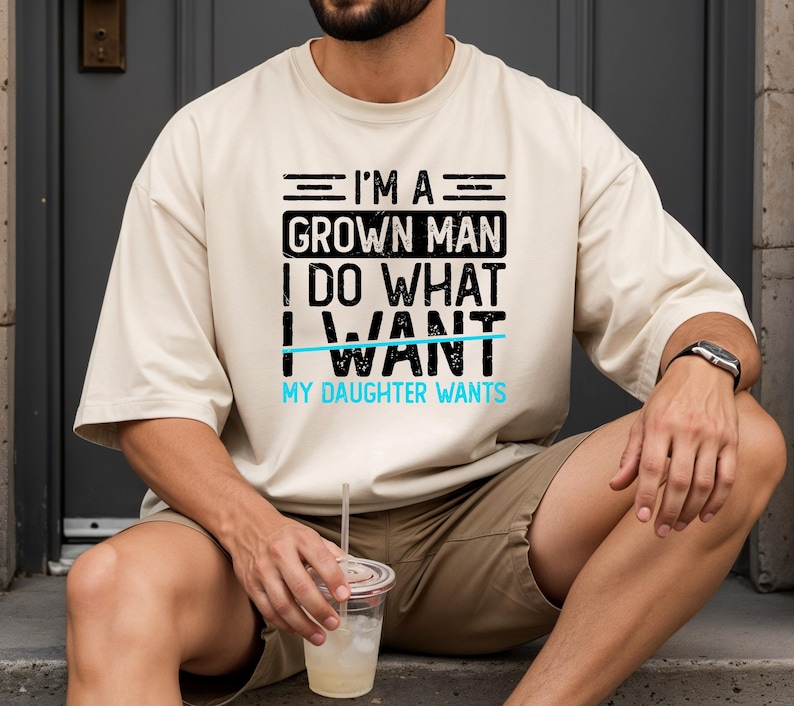 May include: A beige t-shirt with the text "I'M A GROWN MAN I DO WHAT I WANT MY DAUGHTER WANTS" in black and light blue. The person is wearing a watch and holding a drink.