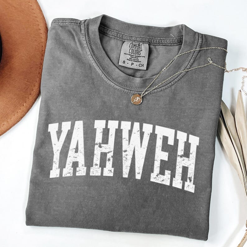 May include: A gray Comfort Colors t-shirt with the word "YAHWEH" in white block letters. A silver necklace with a small pendant rests on the shirt. A brown hat is partially visible in the upper left corner.