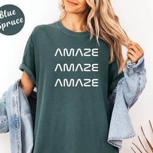 Pu&ograve; includere: Una maglietta color Blue Spruce con la parola "AMAZE" stampata tre volte in bianco. La maglietta ha una vestibilit&agrave; ampia ed &egrave; realizzata in un materiale morbido. La modella indossa una giacca di jeans.